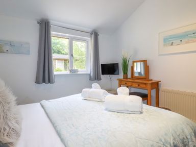A bedroom with a bed, rolled towels on the bed, a window with curtains, a wall-mounted TV, a wooden dressing table with a mirror and a plant at 3 Lake View in Lanreath