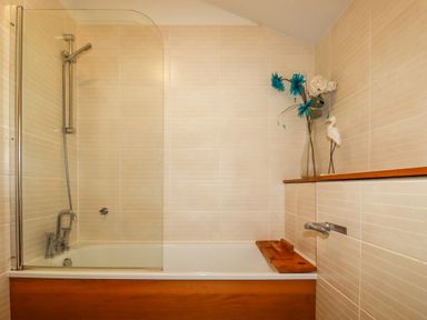 A bathroom with a bathtub shower combination wooden bath panel and shelf with decorative flowers at 3 Lake View in Lanreath