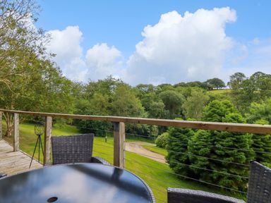 A balcony with wicker chairs and a round table overlooking a green landscape with trees and a pathway at 3 Lake View in Lanreath