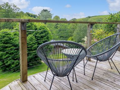An outdoor wooden deck with two black wire chairs and a small round table overlooking green trees and hills at 3 Lake View in Lanreath