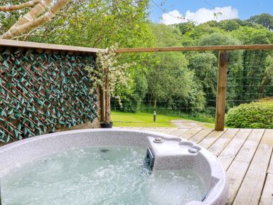 An outdoor hot tub on a wooden deck with green trees and a lawn in the background at 3 Lake View in Lanreath