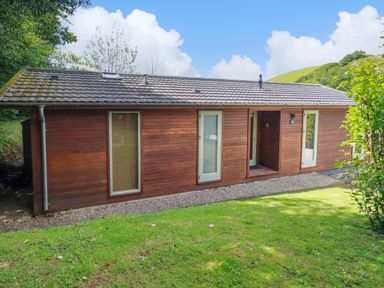 A wooden house with three narrow windows and a small porch surrounded by grass and trees at 1 Hedgerows in Lanreath
