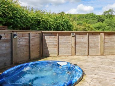 A wooden deck with a blue hot tub and wooden fencing surrounded by green trees at 1 Hedgerows in Lanreath