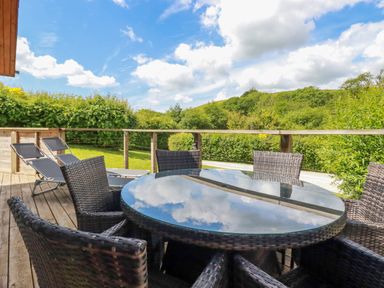 An outdoor wooden deck with a glass top round table and wicker chairs with lounge chairs and greenery in the background at 1 Hedgerows in Lanreath