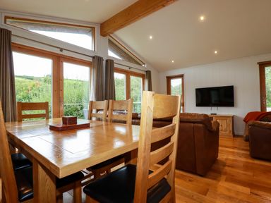 A dining area with wooden chairs and table adjacent to a living room with brown leather sofas and a wall-mounted TV at 1 Hedgerows in Lanreath