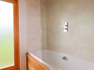 A bathroom with a bathtub that has wooden paneling and beige tiled walls at 1 Hedgerows in Lanreath