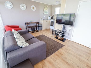 A living room with a gray sofa pillows a red chair tv on a stand a small dining table and kitchenette at 68 Cherry Park in Chapel St Leonards