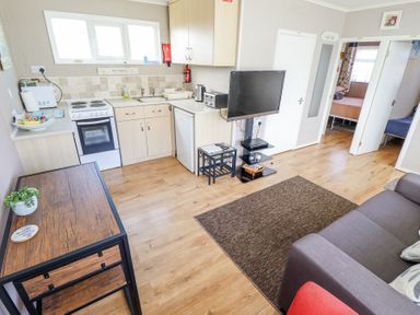 A small kitchen area with stove and refrigerator next to a living room with a couch and TV at 68 Cherry Park in Chapel St Leonards