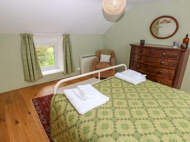 A bedroom with a bed covered in a green patterned blanket and folded towels a wooden chest of drawers with an oval mirror a brown armchair near a small window with green curtains at 2 Summerhill in Letterston