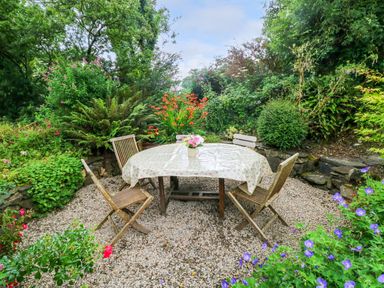 A garden seating area with a table covered by a floral cloth and wooden chairs surrounded by plants and flowers at 2 Summerhill in Letterston