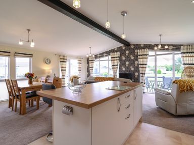 An open plan room with a kitchen island dining table with chairs and living area with sofas and large windows near Owls Retreat Lodge 11 Near Aldeburgh