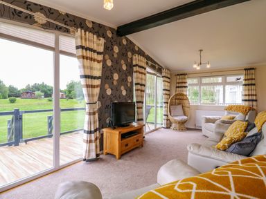 A living room with beige sofas and a wooden TV stand near large windows with striped curtains at Owls Retreat Lodge 11 Near Aldeburgh