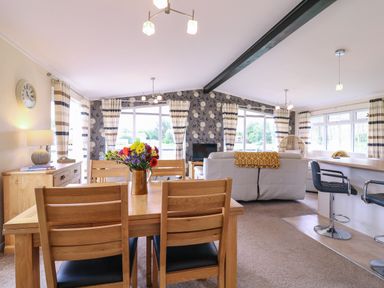 A living room and dining area with wooden furniture flowers on the table and striped curtains at Owls Retreat Lodge 11 Near Aldeburgh