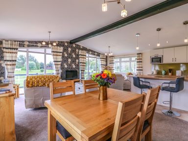 A dining area with wooden table and chairs adjacent to a living room with sofas and a TV and an open kitchen with bar stools at Owls Retreat Lodge 11 Near Aldeburgh
