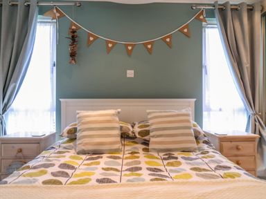 A bedroom with a bed having patterned bedding and two striped pillows between two bedside tables with curtains on windows at Owls Retreat Lodge 11 Near Aldeburgh