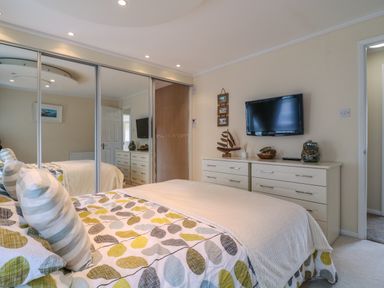 A bedroom with a bed covered in leaf patterned bedding a dresser with decorative items a wall-mounted television and mirrored sliding closet doors at Owls Retreat Lodge 11 Near Aldeburgh