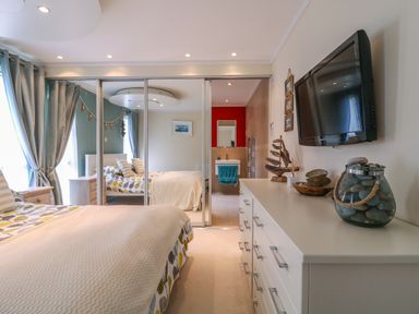 A bedroom with a bed beside windows with curtains a white dresser with decor and a wall-mounted TV and mirrored sliding closet doors reflecting a bathroom at Owls Retreat Lodge 11 Near Aldeburgh