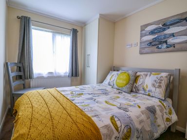 A bedroom with a bed covered in patterned bedding and a yellow blanket near a window with grey curtains at Owls Retreat, Lodge 11 Near Aldeburgh
