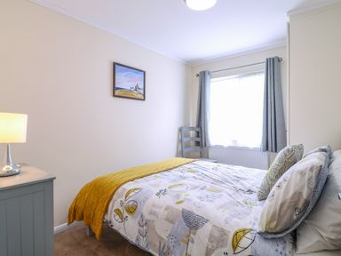 A bedroom with a bed near a window with curtains a bedside table with a lamp a chair and a painting on the wall at Owls Retreat Lodge 11 Near Aldeburgh