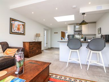 An open living room and kitchen area with a wooden coffee table a brown leather sofa a wooden chest of drawers a kitchen island with two gray bar stools and a refrigerator at 3 Palms in Preston Sands
