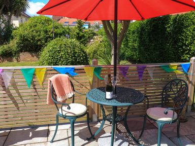A patio set with two chairs and a table under a red umbrella with a bottle and glasses on it and a garden background at 3 Palms in Preston Sands