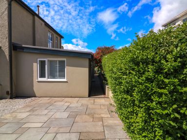 An exterior view of a house with a paved patio area a window with shutters on a wall and a green hedge along a narrow pathway at 3 Palms in Preston Sands