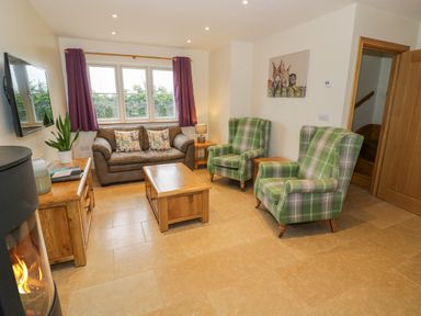 A living room with a brown sofa and two green plaid armchairs a wooden coffee table and side tables a wall mounted television and a wood burning stove at Lavender Cottage in Oxhill near Tysoe