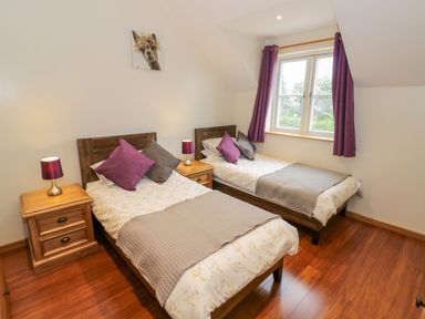 A bedroom with two single beds with pillows and blankets a window with purple curtains and wooden nightstands with lamps at Lavender Cottage in Oxhill near Tysoe