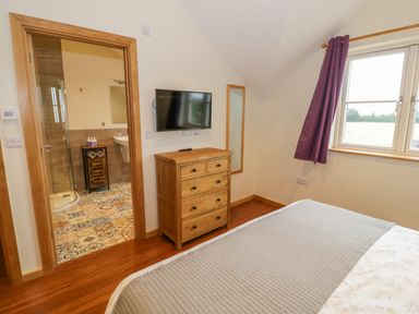 A bedroom with a bed a wooden chest of drawers a wall-mounted TV a mirror a window with curtains and an open door to a bathroom at Lavender Cottage Oxhill near Tysoe