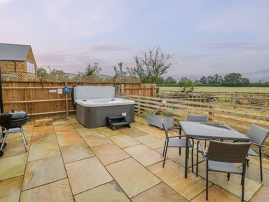 An outdoor patio with a hot tub a barbecue grill a table and chairs and a wooden fence overlooking a field at Lavender Cottage in Oxhill near Tysoe