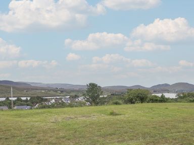 A grassy field with trees and houses in the distance with hills and clouds in the sky at ARD NA GHAOITHE in Loughanure County Donegal