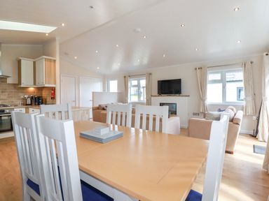 A dining table with white chairs in an open kitchen and living room with sofas and a wall-mounted TV at 43 The Maples in Sticker