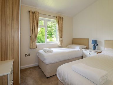 A bedroom with two single beds a window between beige curtains a nightstand with a blue lamp and a wardrobe at 43 The Maples in Sticker
