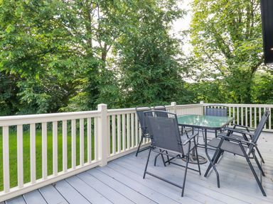 An outdoor deck with a glass table and six black chairs surrounded by trees at 43 The Maples in Sticker