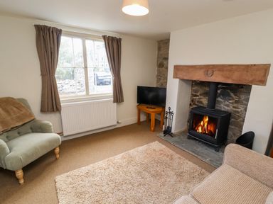 A living room with an armchair, a sofa, a wood burning stove, a small table with a television, and a large window with curtains at Holborn Cottage, Blencathra Street in Keswick