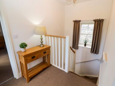 A staircase landing with a wooden side table holding a lamp and a plant next to a window with curtains at Holborn Cottage Blencathra Street Keswick