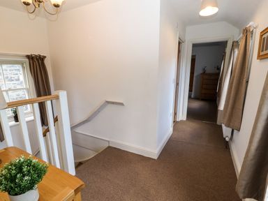 A hallway with brown carpet a wooden table with a plant stairs window with curtains and a room with a wooden chest of drawers at Holborn Cottage Blencathra Street Keswick