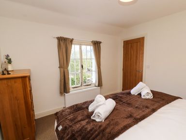 A bedroom with a bed covered by a brown blanket and white towels on it a wooden dresser with small decorations a window with brown curtains and a wooden door at Holborn Cottage Blencathra Street Keswick