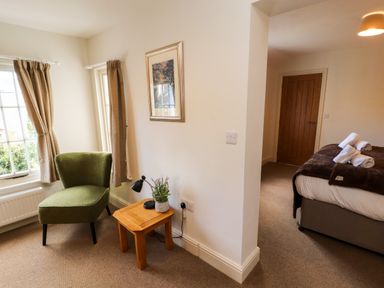 A corner with a green chair and a wooden side table with a lamp and plant next to a bedroom with a bed and rolled towels at Holborn Cottage Blencathra Street in Keswick