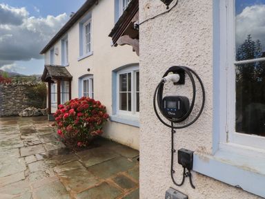 The exterior of a house with a stone patio a red flowering bush and an electric vehicle charging point on the wall at Holborn Cottage Blencathra Street Keswick