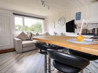 A living area with a gray sofa with cushions a wooden table with black chairs a wall clock a mounted television and a window with a view at Villa 18 Lanteglos Holiday Park in Lanteglos Holiday Park near Camelford