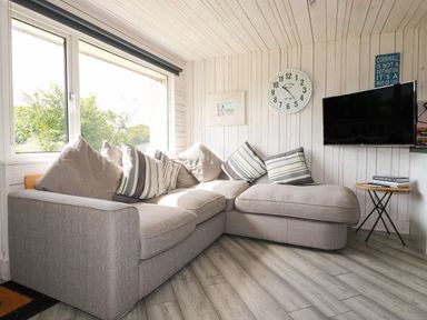A living room with a sectional sofa and cushions a wall clock a TV and a side table with books at Villa 18 Lanteglos Holiday Park near Camelford