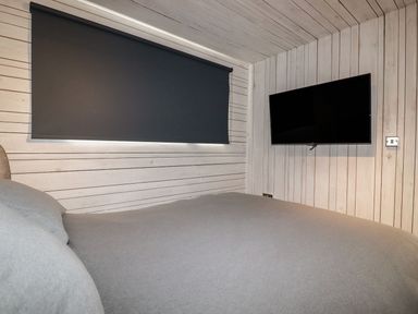 A bedroom with a grey bedspread a wall-mounted television and a closed black window blind at Villa 18 Lanteglos Holiday Park near Camelford