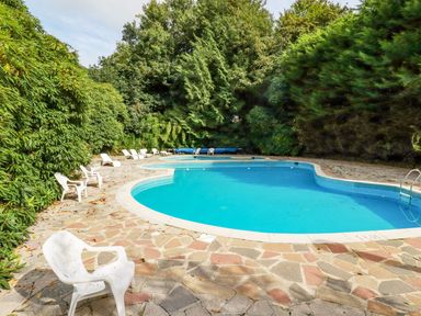 An outdoor swimming pool surrounded by stone paving and white plastic chairs with dense green trees in the background at Villa 18 Lanteglos Holiday Park in Lanteglos Holiday Park near Camelford
