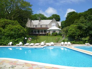 A swimming pool with white lounge chairs and a stone building with a glass conservatory in the background at Villa 18 Lanteglos Holiday Park near Camelford