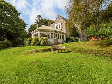 An old stone house with a glass conservatory surrounded by green grass and trees with outdoor seating near a wooden bench and table with chairs at Villa 18 Lanteglos Holiday Park in Lanteglos Holiday Park near Camelford