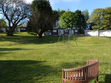 A grassy outdoor area with a curved wooden bench and a swing set near small buildings at Villa 18 Lanteglos Holiday Park in Lanteglos Holiday Park near Camelford
