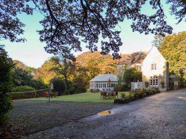 An outdoor area with tables and chairs on grass near a stone building and a white gazebo surrounded by trees at Villa 18 Lanteglos Holiday Park near Camelford