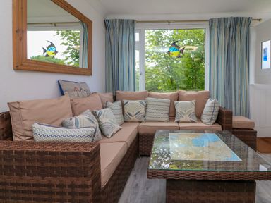 A living room with a brown wicker L shaped sofa with beige cushions and patterned pillows a glass coffee table striped curtains and a window with a fish decoration at 4 The Park in Kilkhampton