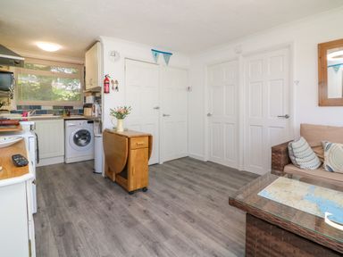 An open kitchen and living area with white doors a washing machine a small wooden foldable table and a wicker sofa with cushions at 4 The Park in Kilkhampton